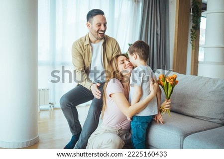 Smiling woman looking at adorable son while sitting on floor with mothers day card and flowers. Adorable kid boy congratulate mom with mother day or birthday embracing mum presenting greeting card