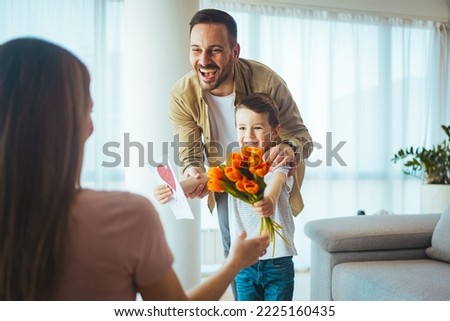  Boy with bunch of beautiful flowers behind back preparing nice surprise for his mother. Cute boy offering flowers and card to his mother in the living room. Handmade Greeting Card For Mother's Day. 