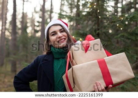 Happy young woman in jumper and coat in santa claus hat with Christmas gifts in  forest. New Year celebration concept greeting card