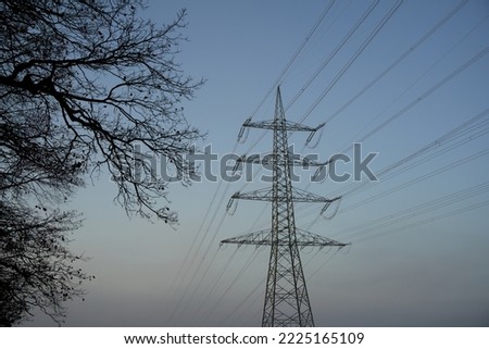 Tall Electricity pylon (Strommasten) also overhead line pylon in foggy Winter morning. Branches from many trees in foreground. Front view.