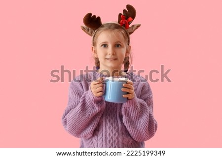 Happy little girl in reindeer horns with cup of cocoa on pink background