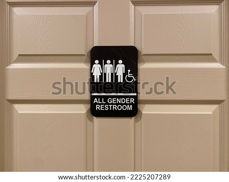 All-gender restroom signage next to a wooden restroom door showing icons of man, woman, transgender and wheelchair user; white icons on black background