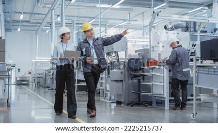 Wide Shot: Two Heavy Industry Employees in Hard Hats Discussing Job Assignments at the Factory, Using Laptop Computer. Asian Engineer and Technician at Work Smiling Royalty-Free Stock Photo #2225215737