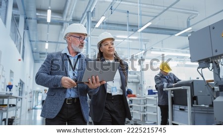Two Professional Heavy Industry Engineers Wearing Hard Hats at Factory. Walking and Discussing Industrial Machine Facility, Working on Laptop. African American Manager and Technician at Work. Royalty-Free Stock Photo #2225215745