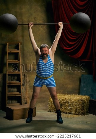 Retro circus. Cinematic portrait of retro circus strongman wearing striped sports swimsuit holding barbell on dark circus backstage background. Concept of creative art, fashion, style and inspiration Royalty-Free Stock Photo #2225217681