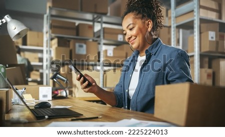 Inventory Manager Using Smartphone to Scan a Barcode on Parcel, Preparing a Small Cardboard Box for Postage. Black Multiethnic Small Business Owner Working on Laptop in Warehouse. Royalty-Free Stock Photo #2225224013
