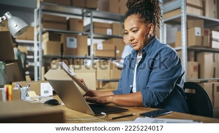 Inventory Manager Using Laptop Computer to Check Order Number on a Parcel, Preparing a Small Cardboard Box for Postage. Shelves Full of Box Packages and Two Colleagues in the Background. Royalty-Free Stock Photo #2225224017