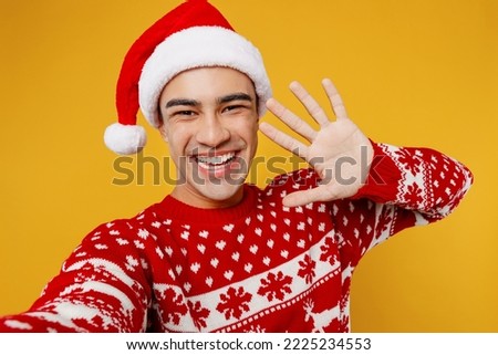 Close up merry young man wear red knitted christmas sweater Santa hat posing do selfie shot pov on mobile cell phone waving hand isolated on plain yellow background Happy New Year 2023 holiday concept