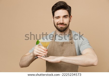 Young cool happy fun man barista barman employee wear brown apron work in bar pub club hold martini glass cocktails wink isolated on plain pastel light beige background. Small business startup concept Royalty-Free Stock Photo #2225234573