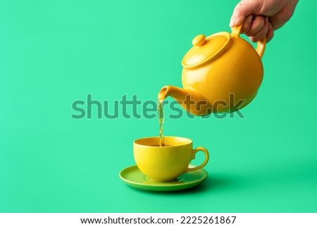 Woman's hand pour tea from yellow jug in a cup, minimalist on a green table. Hot healthy drink, mint tea in a yellow-colored cup. Royalty-Free Stock Photo #2225261867
