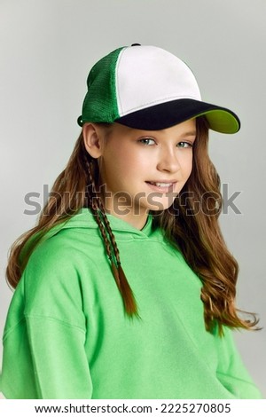 Girl in baseball cap and green hoodie looks in camera smiling. Portrait of teen model with long brown hair posing in studio on grey background closeup