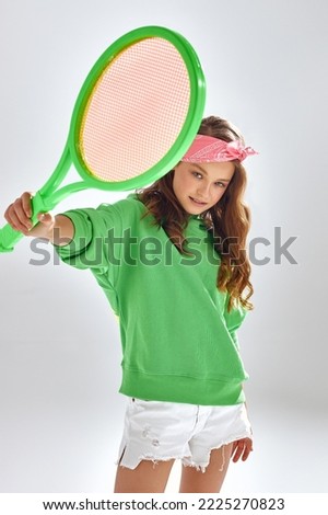 Teen girl with pink headband shows tennis racquet standing in studio. Sportive teenager in green hoodie poses for photo on white background closeup