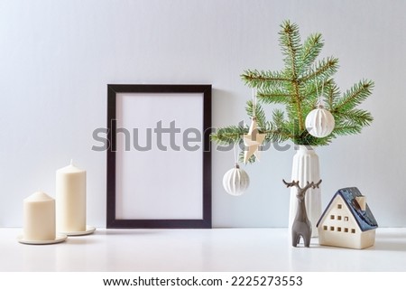 Mock up black poster frame with christmas decoration in home interior, scandinavian style. Green fir branches in a vase, deer and ball on a white table