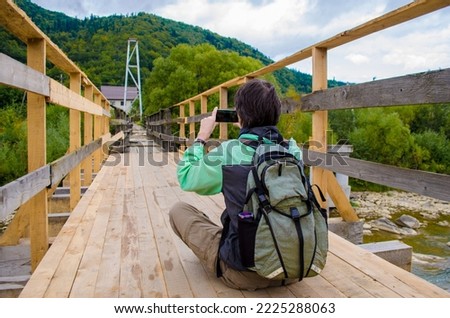 Young man tourist in a green jacket and with a backpack sits on a wooden bridge near the mountains, and takes pictures of them on a smartphone