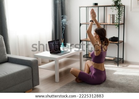 Caucasian young woman practicing yoga in lotus position with namaste gesture at cozy living room. Fitness dark haired lady sitting on floor and using modern laptop.