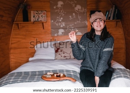 blue-eyed caucasian girl in woolen sweater and brown hat happily smiling at camera sitting on cozy bed in log cabin before eating snack of small cereal toast and cheese, te wepu pods akaroa, new