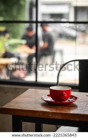 empty cappuccino coffee ceramic mug on saucer on wooden table inside coffee shop against blurred garden with some teenagers. modern lifestyle with coffee brewing