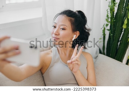 Athletic female Asian blogger filming herself on her phone camera sitting on the couch and smiling with headphones waving, freelance work from home, recording sports workout lifestyle