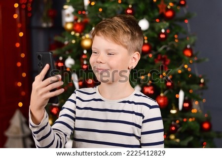 Christmas eve - smiling boy talking photo of themselves on mobile phone. Christmas tree with lights on dark red as background.