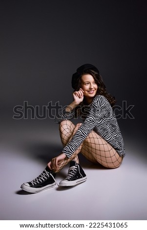 Cheerful woman in fishnet tights sits on floor in studio smiling. Young brunette model enjoys posing for photoshoot on dark grey background