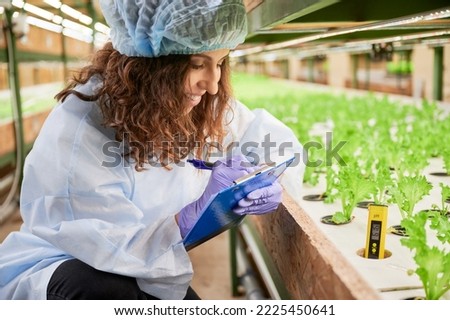 Smiling young woman in rubber garden gloves writing on clipboard while studying plant growth. Female gardener doing research and filling out report in greenhouse.