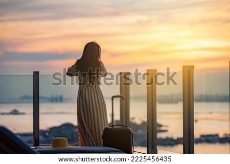 Beautiful woman in dress with suitcase on vacation by tropical beach, standing on the glass balcony enjoy fresha air with ocean view. Golden sunset sky on the beach, Beautiful cloudscape over the sea. Royalty-Free Stock Photo #2225456435