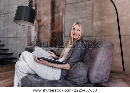 Gorgeous blonde fashion model relaxing at home, sitting with book on cozy chair looks aside happily  against modern interior. Successful businesswoman relaxing after hard week. Purposeful female. Royalty-Free Stock Photo #2225457653