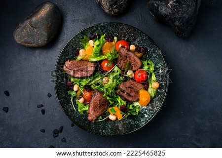 Fresh salads with beef steak, tomatoes, green leaves, arugula, tangerines and hazelnuts on a dark background. Healthy food, clean eating. Salad beef steaks, lettuce, arugula. Top view. Royalty-Free Stock Photo #2225465825
