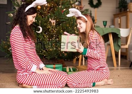 Daughter giving a gift box to her mother near christmas tree. Happy family wearing striped pajamas and rabbit ears celebrating Christmas or New Year of the Rabbit. Selective focus.