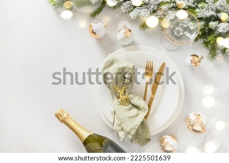 Beautiful white Christmas table setting with golden balls, bottle of champagne and napkin ring as deer on white background. View from above. Copy space. Xmas festive dinner. Royalty-Free Stock Photo #2225501639