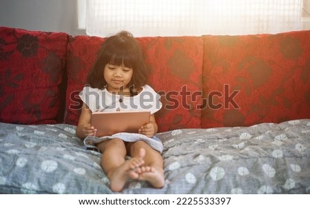 Toddler girl playing with digital wireless tablet computer on couch at home. Kid Having Fun Browsing Internet And Playing Game Online On Computer. Children And Gadgets
