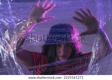 Artistic depiction of overwhelming anxiety. Young attractive long-haired caucasian girl in striped t-shirt trapped behind transparent plastic foil. Violet lightening. High quality photo