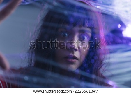 Scared female caucasian teenager with long black hair and bangs looking from behind transparent plastic-like barrier. Depiction of anxiety. Closeup portrait. Makeup gems on face. High quality photo