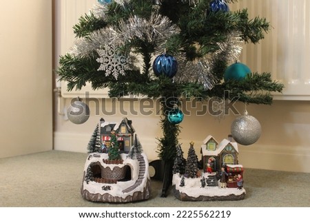 A Miniature Christmas village scene with snow covered houses, a stall, trees and people celebrating the holidays. The models are set underneath a Christmas tree with blue and silver decorations.