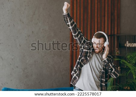 Young happy singer fun cool man 20s he wears brown shirt headphones listen to music sing song dance raise up hand stand near blue sofa in own living room apartment stay home indoor flat on weekends.