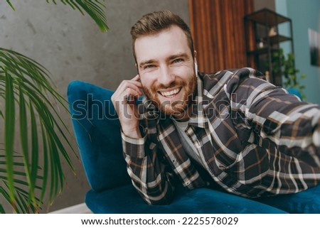 Close up young man 20s he wears brown shirt listen music in earphones doing selfie shot pov on mobile cell phone sitting on blue sofa in own living room apartment stay home indoor flat on weekends.