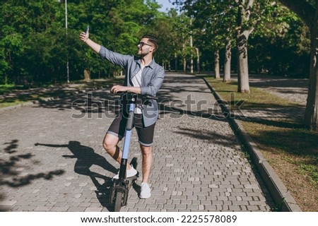 Full length young man in blue shirt shorts glasses do selfie shot on mobile phone riding scooter go down alley rest in spring green city park sunshine lawn outside on nature Urban leisure concept