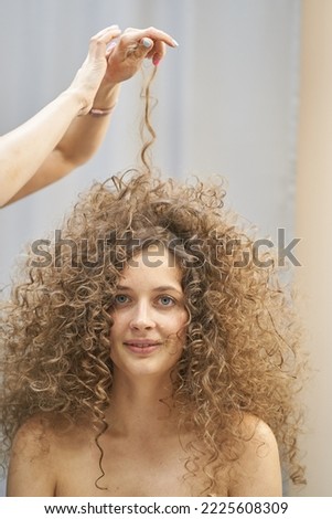 The hair master in face mask makes a curly hairstyle for a smiling woman. Portrait of a beautiful Caucasian girl with flowing curly hair. The concept of beauty salon. High quality photo