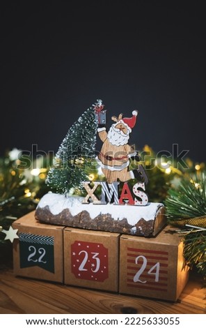 Beautiful three kraft boxes advent calendar with numbers 22,23,24 on a wooden table with christmas decor and a burning garland on a blurred dark background, close-up side view. Royalty-Free Stock Photo #2225633055