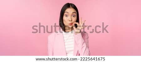 Portrait of asian corporate woman showing mouth seal, close shut lips on key gesture, promise keep secret, standing over pink background in suit. Royalty-Free Stock Photo #2225641675