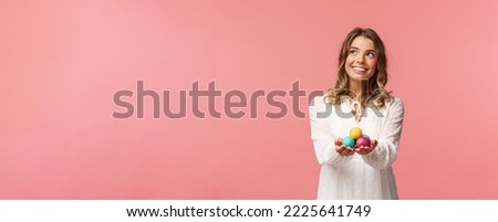 Holidays, spring and party concept. Portrait of dreamy good-looking blond girl in white dress, holding painted eggs, celebrating Easter, orthodox holy day, smiling and looking away.