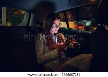 Beautiful business woman using smartphone in a car at night. Busy female working on a mobile phone in the back seat of a taxi. Bokeh and neon lights in the background. Illuminated by the screen device Royalty-Free Stock Photo #2225657381