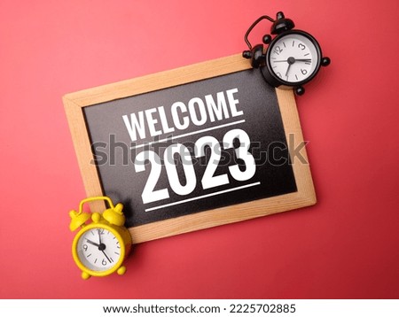 Wooden frame and alarm clock with the word WELCOME 2023 on red background.