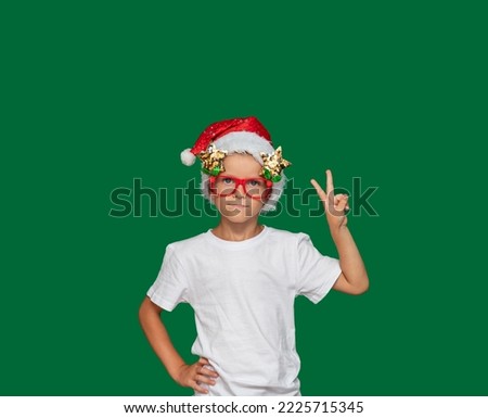 Boy in Santa Claus hat and glasses shows peace sign with his hand. Green background with space for text. Selective focus. Picture for articles and advertisements about children, holidays, Christmas.