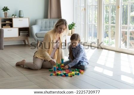 Loving mother play with little 6s daughter at home, using wooden cubes build castle sit together on warm floor in cozy sunny living room. Family pastime and leisure, developmental games with children Royalty-Free Stock Photo #2225720977