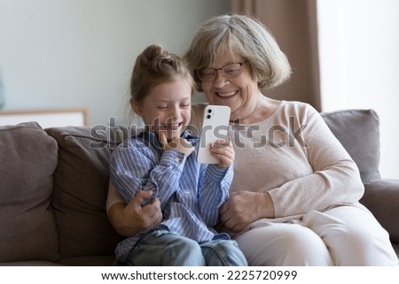 Little girl spend time with older great-grandmother have fun use new mobile application, look at smartphone screen enjoy online amusing app. Multi generational family leisure at home with modern tech Royalty-Free Stock Photo #2225720999
