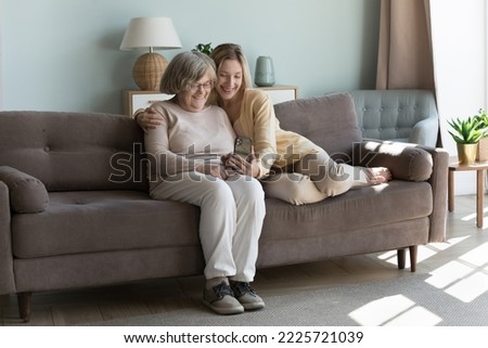 Caring adult granddaughter hugs her elderly grandmother looking together on modern smartphone screen, make video call, take selfie picture spend leisure use wireless tech, internet connection at home
