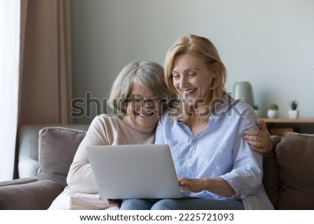 Mature daughter and old mother sit on sofa with laptop choose goods, buy services via internet, web surfing together at home. Younger relative teaching using computer, showing useful apps and websites Royalty-Free Stock Photo #2225721093