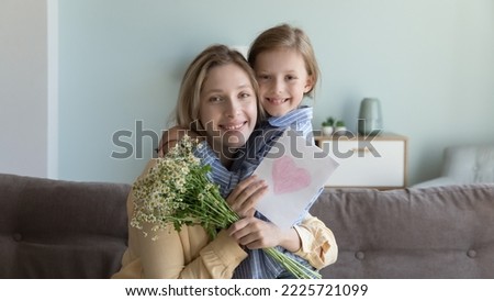 Portrait of smiling young mother and little daughter sitting on couch look at camera celebrating birthday together, loving girl congratulate happy mom with anniversary embracing hold flowers and card