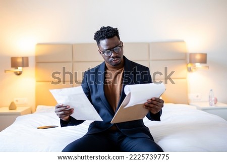 Serious Arican business man sitting on hotel bed working, analyzes expenses on business trip at the hotel room.